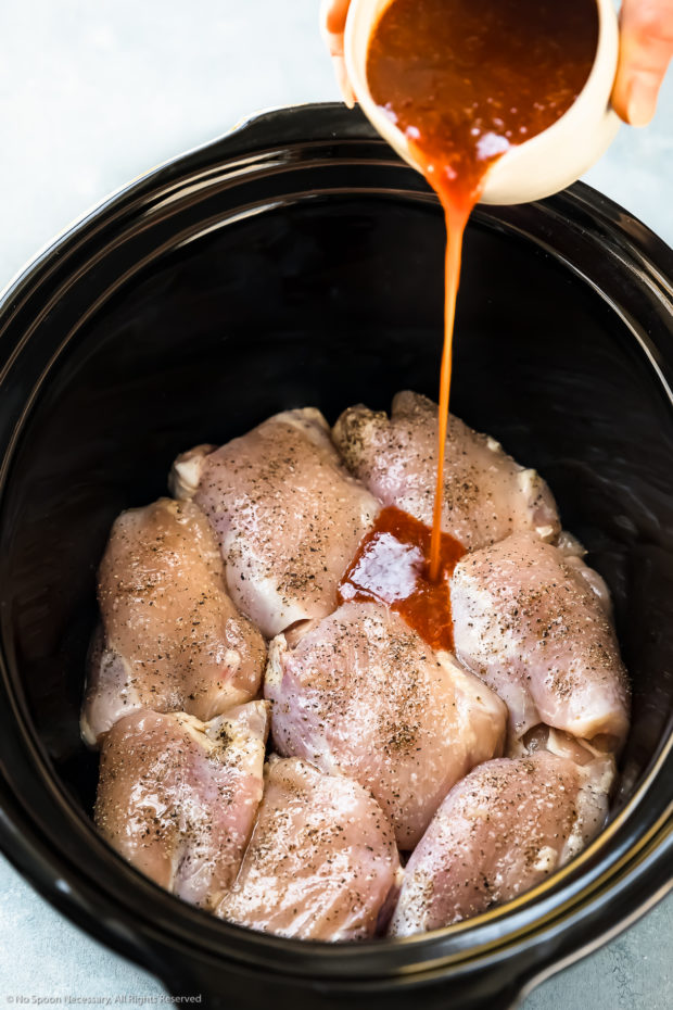 Angled photo of uncooked chicken thighs in the bowl of a slow cooker with Thai sweet chili sauce being poured on top of the chicken - photo of step 2 of the recipe.