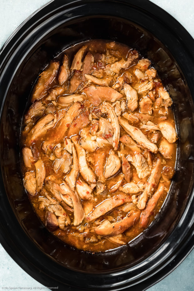 Overhead photo of cooked, shredded chicken thighs coated in a sweet chili sauce in a black slow cooker - photo of stew 4 of the recipe.