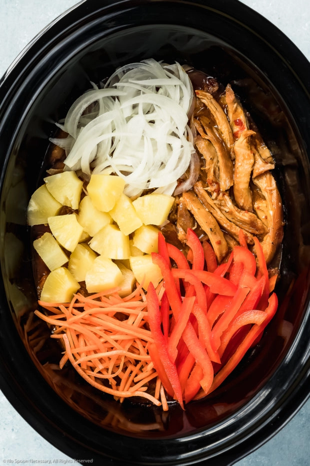 Overhead photo of a black slow cooker filled with sliced onions, bell peppers, carrots, pineapple chunks and cooked, shredded sweet chili chicken - photo of step 5 of the recipe.