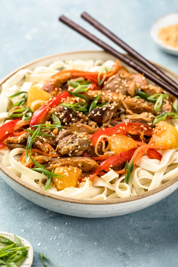 Angled shot of Slow Cooker Sweet Chili Chicken garnished with sesame seeds and sliced scallions on a bed of lo mein noodles in a neutral colored bowl with a pair of wooden chopsticks resting on the side of the bowl.