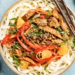 Overhead, landscape shot of Slow Cooker Sweet Chili Chicken on a bed of lo mein noodles in a neutral colored bowl with a pair of wooden chopsticks resting on the side of the bowl and ramekins of sesame seeds and sliced scallions arranged around the bowl.