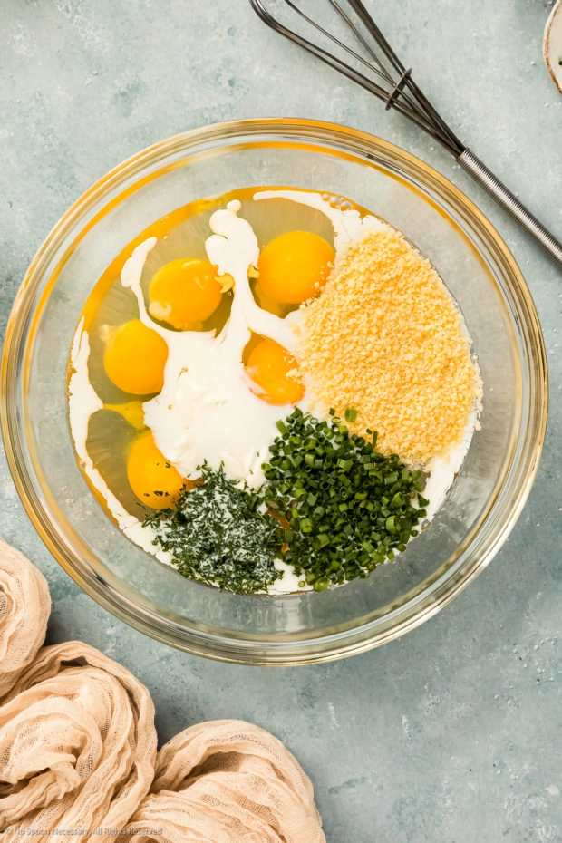 Overhead photo of a glass mixing bowl fill with raw eggs, milk, grated parmesan and fresh herbs. 