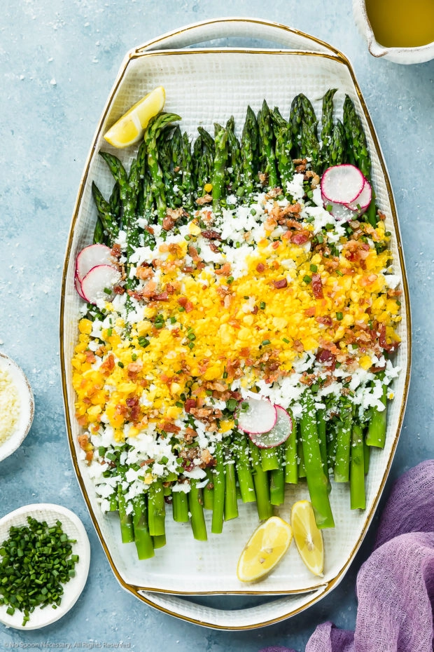 Overhead photo of Asparagus Mimosa garnished with crumbled bacon, grated parmesan and slices of radishes on a large white platter with a mini pouring jug of vinaigrette and ramekins of grated cheese and snipped chives arranged around the platter.