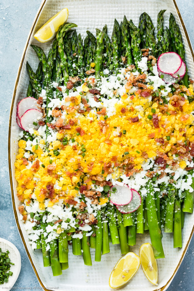 Overhead, up-close photo of Asparagus Mimosa garnished with crumbled bacon, grated parmesan, slices of radishes and lemon wedges on a large white platter with a ramekin of snipped chives tucked in the bottom left corner of the shot.