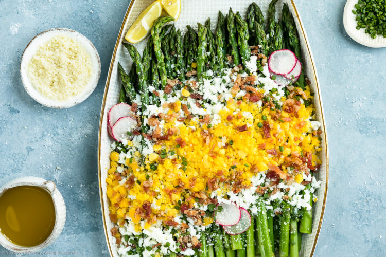 Overhead, landscape photo of Asparagus Mimosa garnished with crumbled bacon, grated parmesan and slices of radishes on a large white platter with a mini pouring jug of vinaigrette and ramekins of grated cheese and snipped chives arranged around the platter.