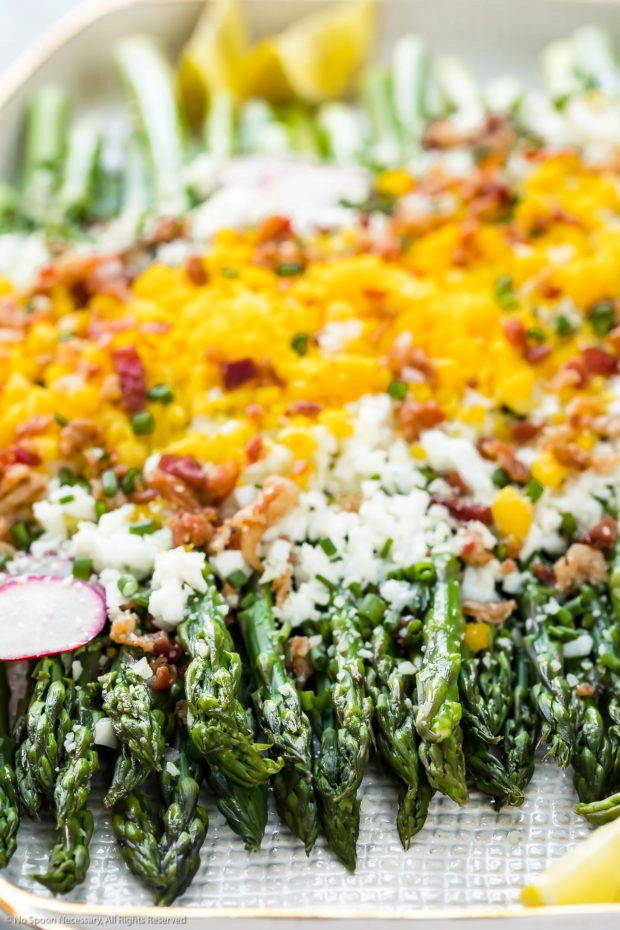 Angled, up-close photo of Asparagus Mimosa garnished with crumbled bacon, grated parmesan and slices of radishes on a large white platter with the focus of the shot on the asparagus spears tips.