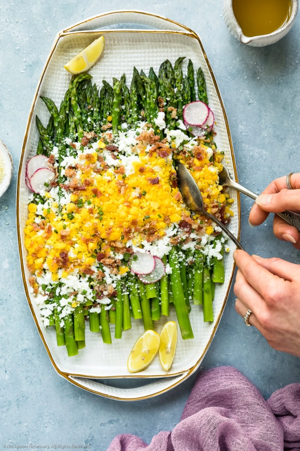 Overhead photo of Asparagus Mimosa garnished with crumbled bacon, grated parmesan and slices of radishes on a large white platter with two hands holding serving utensils tucked under the asparagus to serve it.