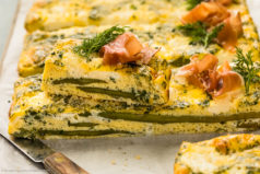 Straight on photo of a slice of eggs and asparagus frittata topped with cheese and herbs.
