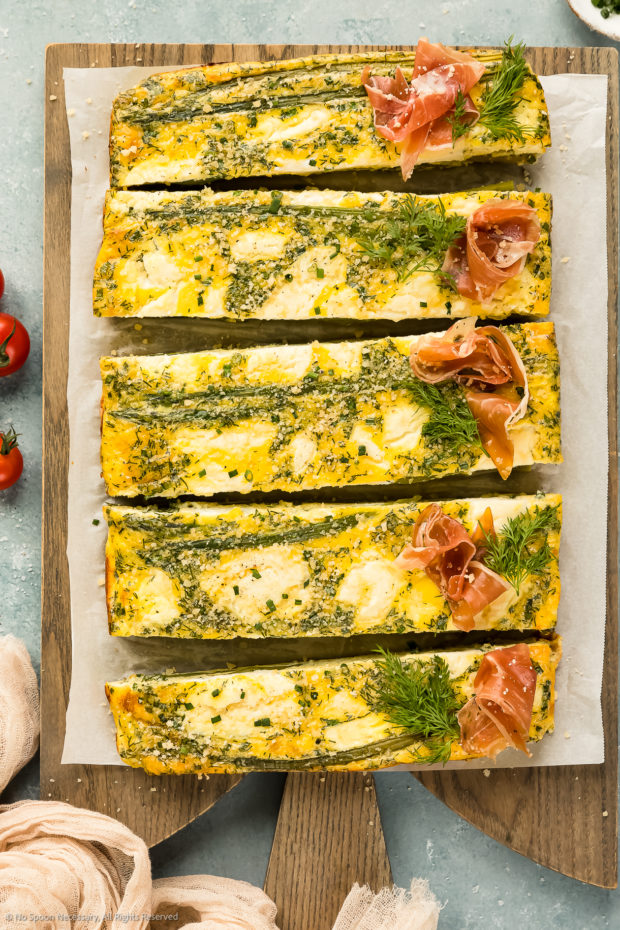 Overhead photo of Asparagus Frittata baked and sliced into long pieces and topped with prosciutto on a parchment paper lined wood serving board.