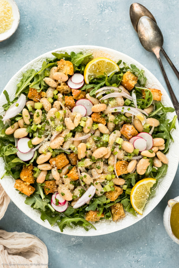 Overhead photo of Cannellini Bean Salad with arugula, radishes and lemon wedges on a white serving platter with a cup of lemon dressing off to the side. 