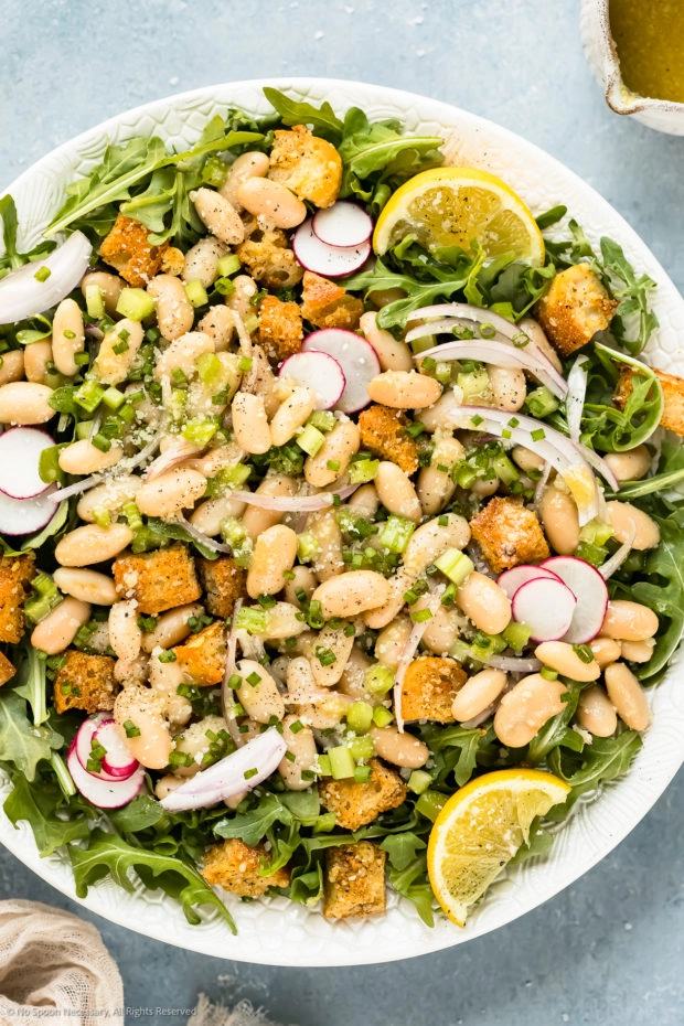 Overhead, close-up photo of marinated beans on a bed of arugula with croutons and lemon wedges. 