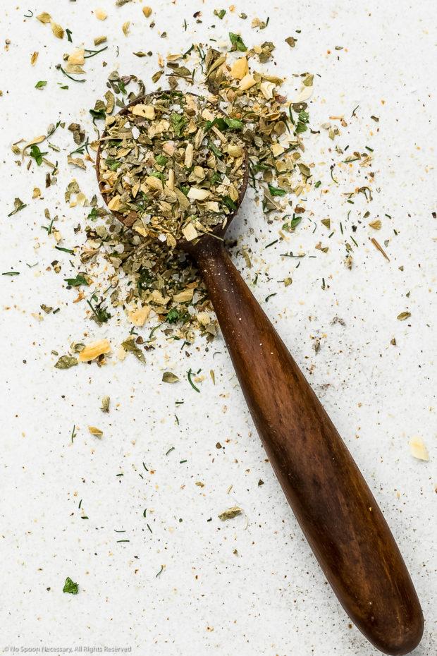 Overhead, up-close shot of a small wooden teaspoon filled with Greek spice blend.