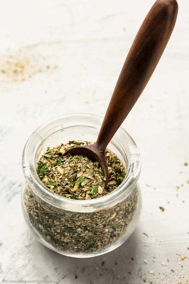 Angled shot of a glass jar filled with homemade greek spices with a small wooden teaspoon inserted into the jar.