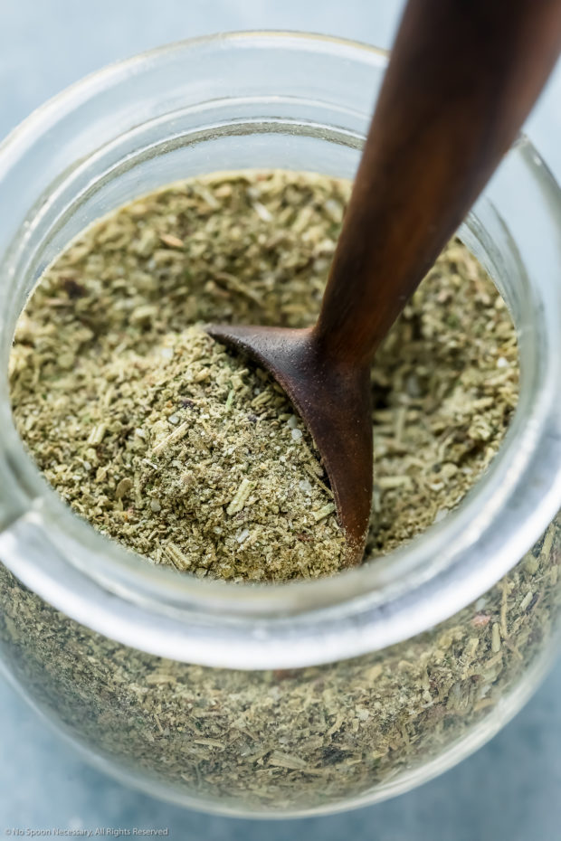 Angled, up-close shot of Italian Seasoning Mix in a small glass jar with a wooden teaspoon inserted into the jar.