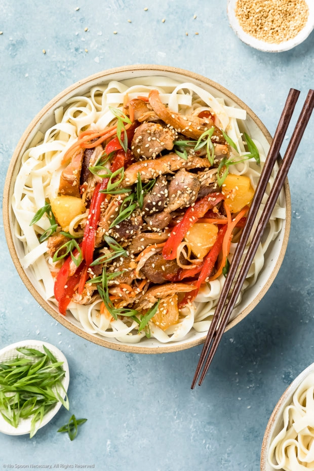 Overhead shot of Slow Cooker Sweet Chili Chicken on a bed of lo mein noodles in a neutral colored bowl with a pair of wooden chopsticks resting on the side of the bowl and ramekins of sesame seeds and sliced scallions arranged around the bowl.