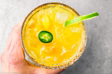 Overhead photo of a hand holding a spicy mango margarita garnished with a jalapeno slice and wheel of lime.