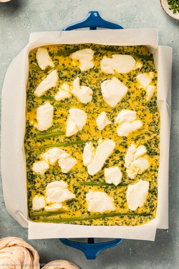 Overhead photo of a parchment paper lined baking dish filled with whisked eggs and asparagus topped with dollops of ricotta (before being baked). 