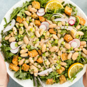 Overhead photo of two hands holding a serving platter of white bean salad over a bed of arugula with lemon wedges.