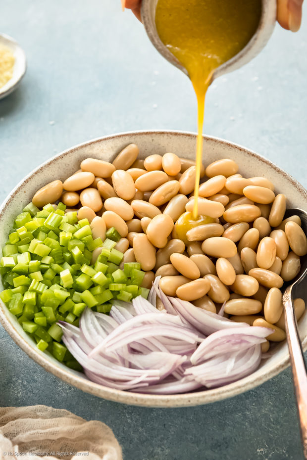 Angled photo of lemon vinaigrette being poured over white beans with chopped celery and sliced scallions in a medium bowl. 