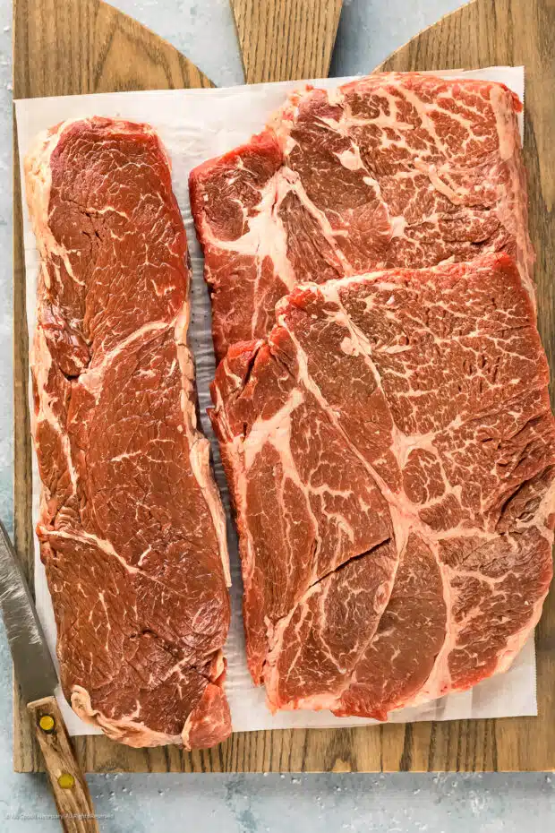 Overhead shot of a gray wood board topped with a sirloin steak and boneless chuck steaks - photo of the two best meat cuts for hamburgers.