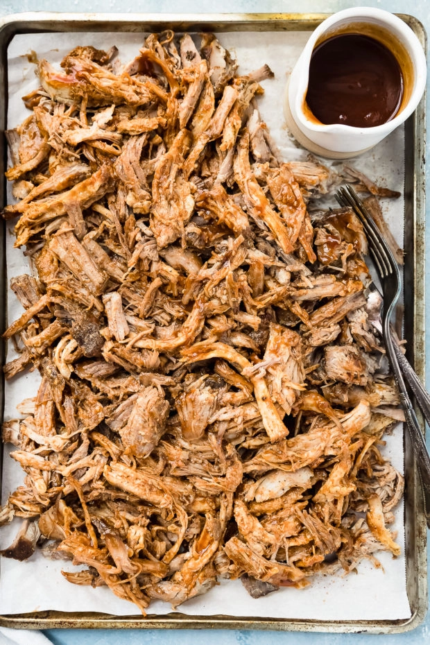 Overhead photo of a parchment paper lined sheet pan topped with pulled pork tossed in Carolina BBQ sauce with a ramekin of more sauce in the corner of the pan.