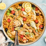 Overhead, landscape photo of Mediterranean Chicken Pasta in a large gray enamel skillet with lemon wedges, a ramekin of fresh oregano and a neutral colored linen arranged around the skillet.