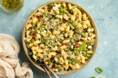 Overhead photo of Bacon Pea Pasta Salad in a white bowl with serving spoons tucked into the salad and a neutral linen, jar of basil vinaigrette and ramekin of fresh basil arranged around the bowl.