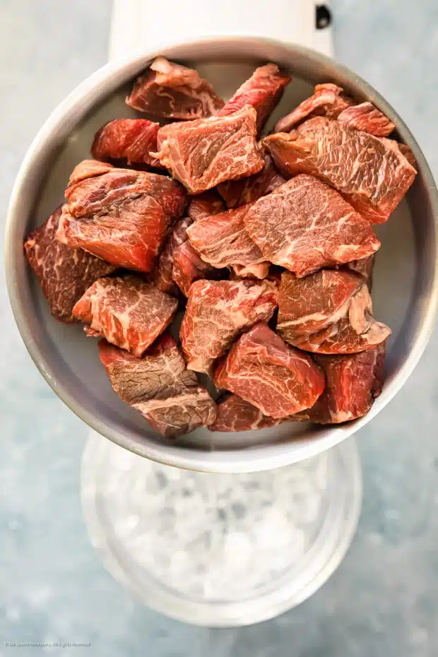 Overhead photo of cubed steak meat for burgers in the tray of the meat grinder hopper with a bowl resting inside an ice water bowl below the grinder.