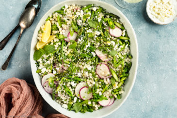 Overhead photo of Lemon Couscous Salad in a neutral colored serving bowl with a ramekin of crumbled ricotta salata, white wine glass, spoons and pink linen arranged around the bowl.