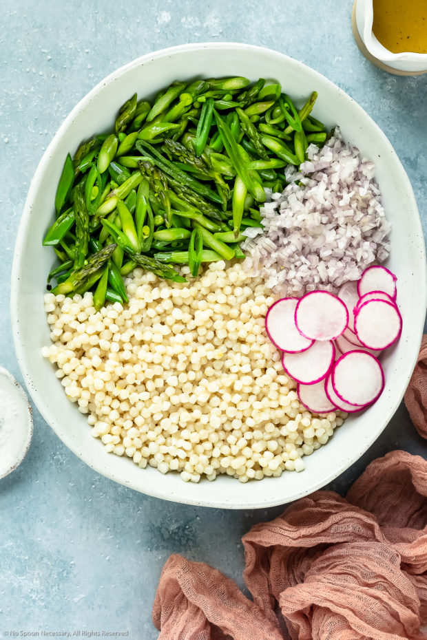 Overhead photo of cooked couscous, sautéed asparagus and snap peas, diced shallots and sliced radishes in a large bowl with a jar of lemon dressing and ramekin of salt arranged around the bowl - photo of step 4 of the lemon couscous salad recipe.