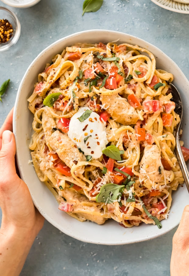 Overhead photo of two hands holding a serving bowl filled with Margarita chicken pasta topped with a dollop of ricotta.