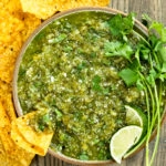 Overhead photo of Salsa Verde topped with lime wedges and fresh cilantro in a neutral colored with yellow tortilla chips scattered around the bowl and a chip inserted into the salsa.