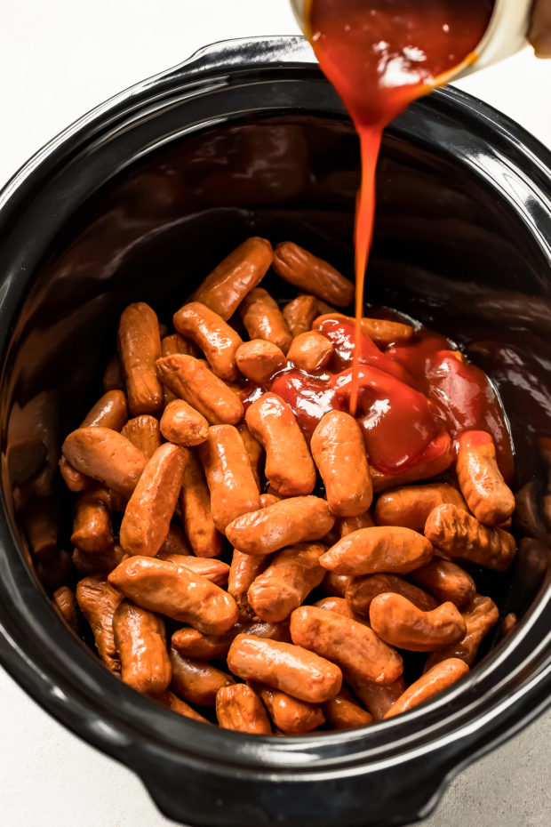 Overhead shot of little smokies covered with honey sriracha sauce in a black slow cooker - photo of the second part of step 2 of the Slow Cooker Honey Sriracha Little Smokies recipe.