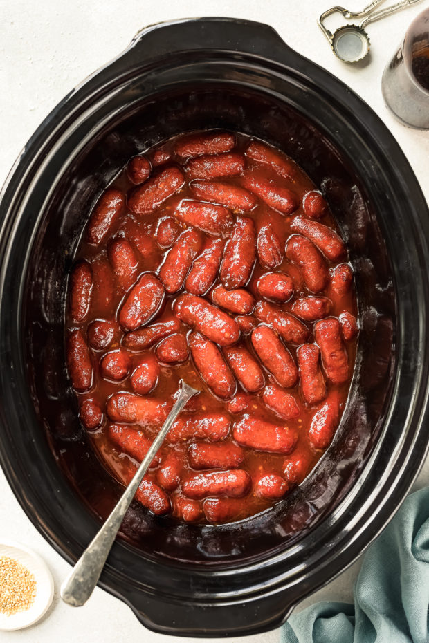 Overhead shot of cooked honey sriracha little smokies in black slow cooker with a spoon inserted into the cooker - photo of step 3 of the Slow Cooker Honey Sriracha Little Smokies recipe.