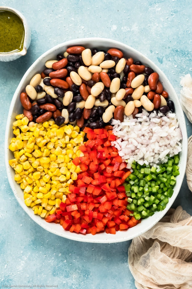 Overhead photo of beans, corn, bell peppers, jalapeno and shallots neatly organized in a large bowl with a jar of cilantro vinaigrette off to the side - step 2 of the Mexican bean salad recipe.