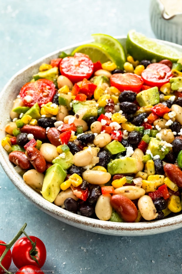 Angled photo of a Mexican Bean salad with avocado and tomatoes in a serving bowl with a small jar of cilantro vinaigrette blurred in the background - step 4 of the Mexican bean salad recipe.