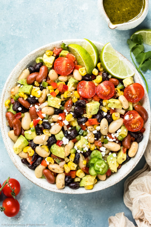 Overhead photo of a Mexican Bean salad garnished with lime wedges in a serving bowl with a small jar of cilantro vinaigrette, vine ripe tomatoes and fresh cilantro arranged around the bowl.