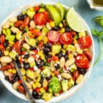 Overhead photo of a Mexican Bean salad with a spoon inserted into the salad in a serving bowl with a small jar of cilantro vinaigrette, vine ripe tomatoes and fresh cilantro arranged around the bowl.