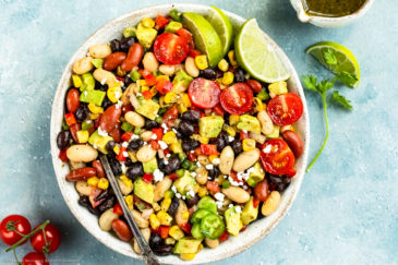Overhead photo of a Mexican Bean salad with a spoon inserted into the salad in a serving bowl with a small jar of cilantro vinaigrette, vine ripe tomatoes and fresh cilantro arranged around the bowl.