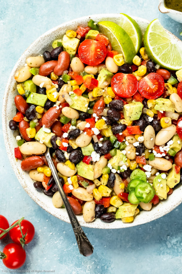 Overhead, up close photo of a Mexican Bean salad garnished with lime wedges in a serving bowl with a spoon inserted into the salad.