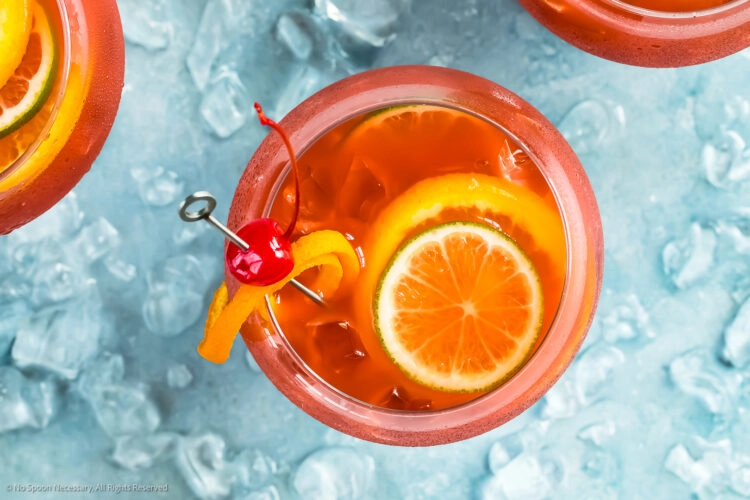 Overhead photo of three Rum Runners garnished with slices of lime and orange with a ramekin of maraschino cherries and a glass shot glass off to the side of the cocktails.