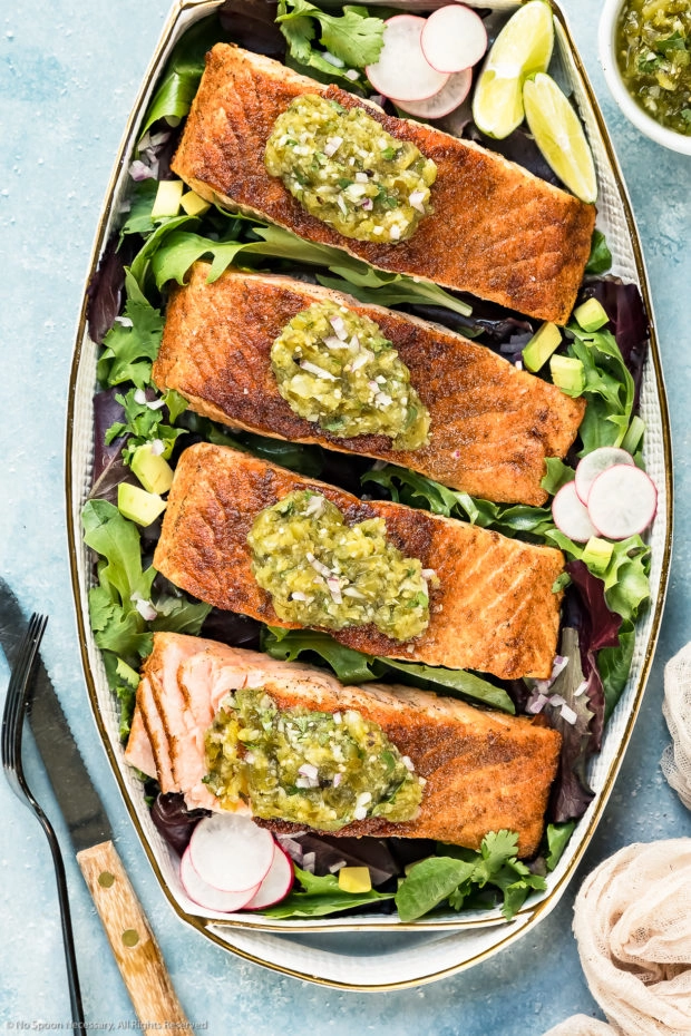 Overhead photo of a white platter topped with pan-seared Salmon garnished with Salsa Verde on a bed of greens with on of the salmon filets cut open to show the inside of the salmon.