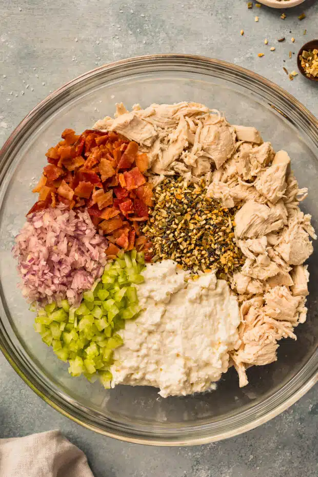Overhead photo of cooked, shredded chicken, cream cheese, crispy bacon, celery, onions, and seasonings in a large mixing bowl.