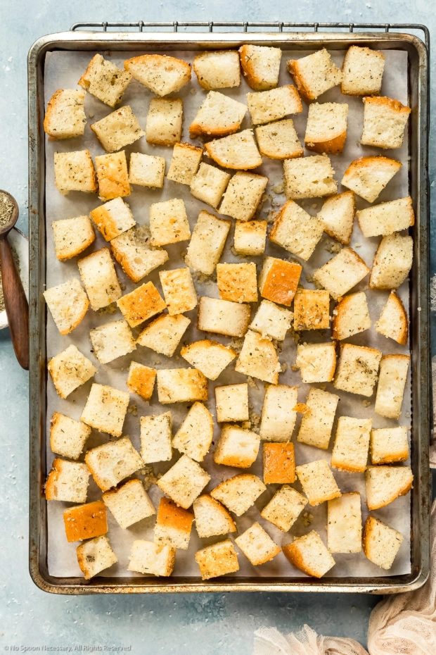 Overhead photo of french bread cubes tossed in oil and seasoning spread out on a parchment paper lined sheet pan - photo of the step 4 of homemade crouton recipe.