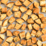 Overhead photo of homemade croutons on a parchment paper lined baking sheet with a ramekin of italian seasoning and neutral colored linen napkin next to the pan.