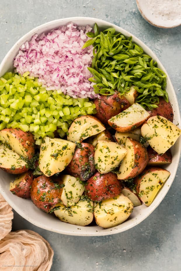 Overhead photo of vinaigrette dressed baby potatoes, chopped celery, chopped red onions, and sliced scallions neatly organized in a mixing bowl.