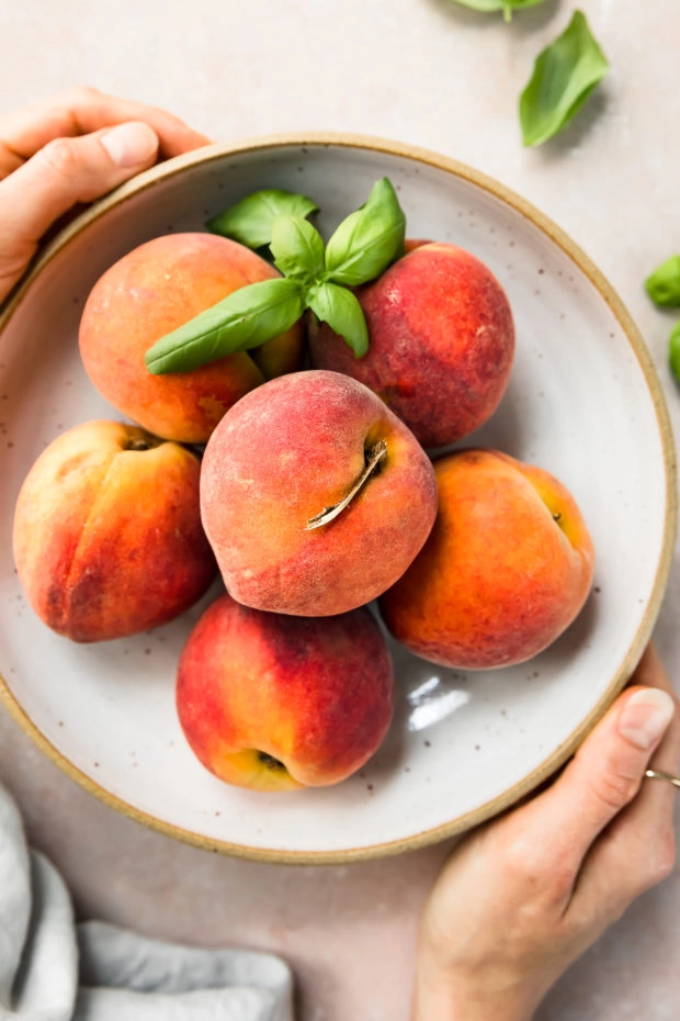 Overhead photo of a white bowl filled with fresh peaches and sprigs of basil with two hands holding the bowl. 