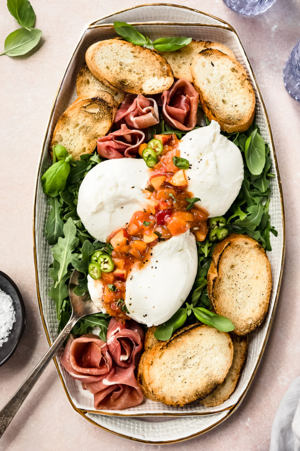 Overhead photo of a white serving platter filled with arugula, burrata, prosciutto, Peach Chutney and toasted slices of baguette.