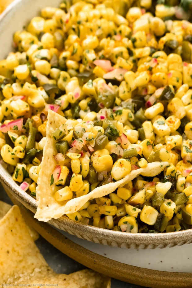 Angled, close-up photo of fresh corn salsa on a tortilla chip in a large serving bowl.