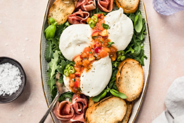 Overhead, landscape photo of a white serving platter with arugula, burrata, prosciutto, Peach Chutney and toasted slices of baguette.